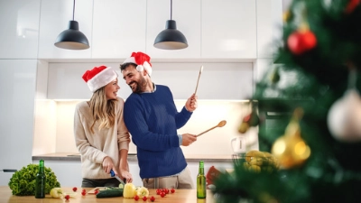 Un couple avec des bonnets de Noël s’amuse en cuisine. L’homme semble jouer de la batterie avec des cuillères en bois.