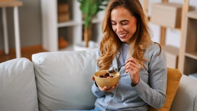Une jeune femme assise sur un canapé mange un bol de céréales en souriant.
