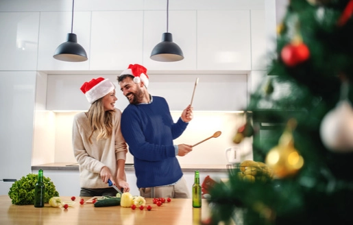 Un couple avec des bonnets de Noël s’amuse en cuisine. L’homme semble jouer de la batterie avec des cuillères en bois.