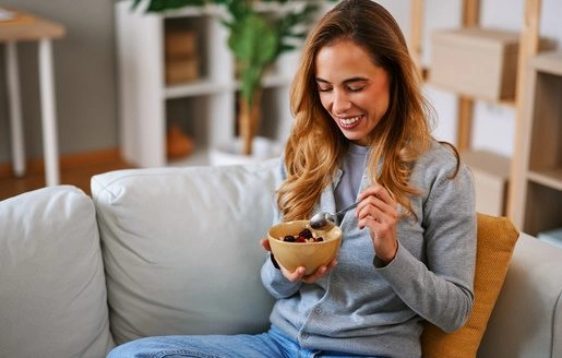 Une jeune femme assise sur un canapé mange un bol de céréales en souriant.