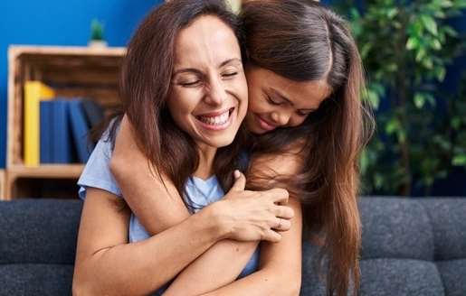 Une petite fille serre sa maman dans ses bras. Elle sourient toutes les deux.