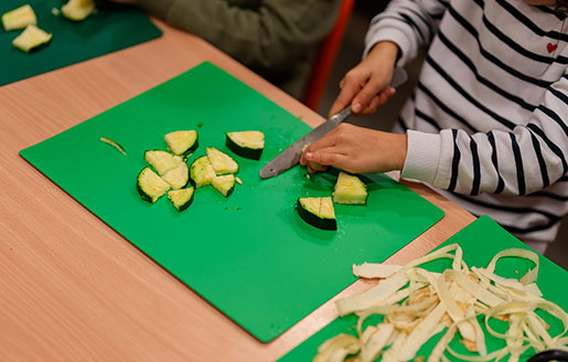 Une jeune enfant découpe attentivement une courgette sur une planche à découper.